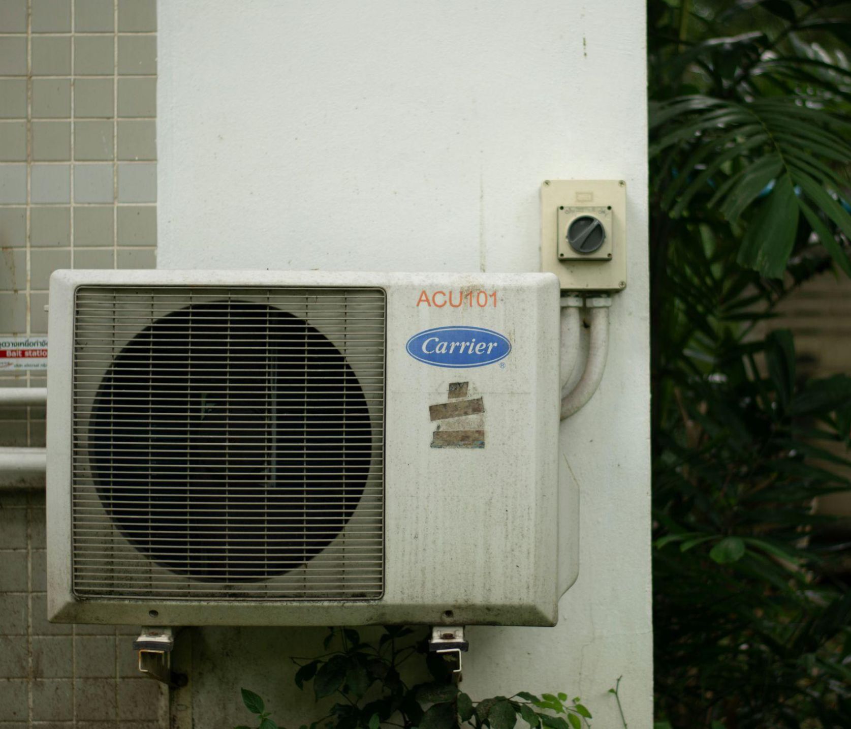 A wall-mounted air conditioning unit surrounded by greenery shows an outdoor cooling system that may require regular maintenance.
